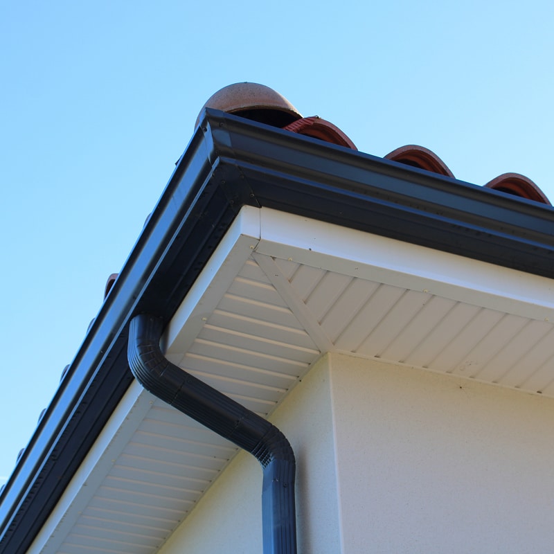 Gouttière noire moderne et descente pluviale Angle de toit moderne avec gouttière noire et descente pluviale. Soffite blanc et tuiles méditerranéennes sous ciel bleu.